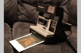 Black and white Polaroid film camera placed on a dark couch with a film photo partially hanging out